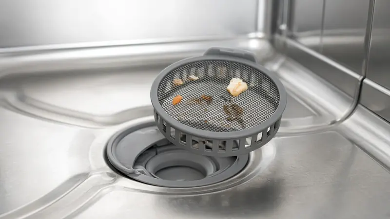 A dishwasher filter cup lifted from the tub floor, held above the open machine